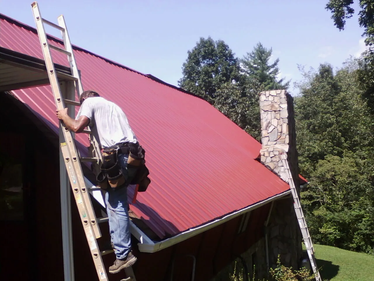 Roofer on a ladder beside a red metal roof and stone chimney — Soffit & Fascia Repair by Roofing Brothers in Hammonton
